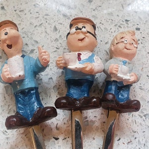 May include: Three ceramic figurines of men with painted facial features, wearing blue and white clothing, holding cups. The figurines are attached to metal sticks.