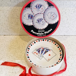 Puede incluir: Un juego de cuatro platos de porcelana, cada uno de 16,5 cm de diámetro, con diseños de naipes. Los platos son blancos con detalles en rojo, negro y azul, y están empaquetados en una caja redonda con una cinta roja.