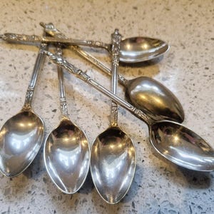 Puede incluir: Una colección de cucharas de plata ornamentadas con diseños detallados en los mangos. Las cucharas están dispuestas de forma ligeramente superpuesta, reflejando la luz y creando reflejos en sus superficies pulidas. Las cucharas son probablemente para servir o comer.