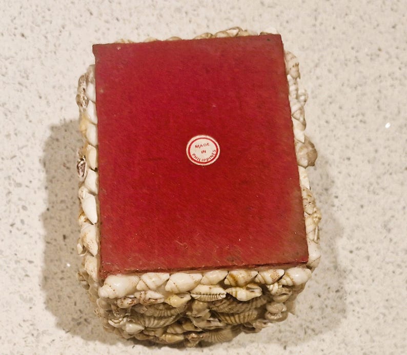 May include: A rectangular box decorated with small, white seashells. The top is a solid, deep red colour. A small, circular sticker on the top reads "MADE IN PHILIPPINES".