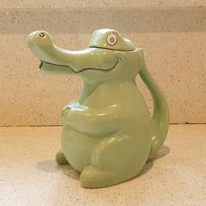 以下が含まれることがあります： 笑顔と片目を持つワニの形をした緑色のセラミック製のティーポット。ティーポットには蓋と注ぎ口があります。