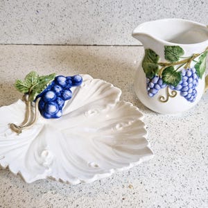 Peut inclure: Une cruche et un plat en forme de feuille en céramique blanche avec des motifs de grappes de raisin. La cruche a une anse en forme de bambou et des motifs de vigne. Le plat présente une grappe de raisins bleus et de feuilles vertes.