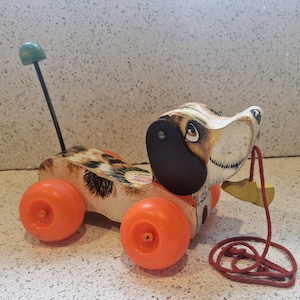 Op de afbeelding: Vintage Fisher-Price trek-hondenspeelgoed. De houten speelgoedhond heeft een gevlekt lichaam, oranje wielen en een zwarte neus. Het heeft een rood trekkoord en een groene handgreep. Een klassiek kinderspeelgoed.
