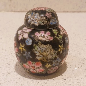 Op de afbeelding: Een kleine, ronde, zwarte porseleinen pot met een deksel. De pot is versierd met een kleurrijk bloemenpatroon.