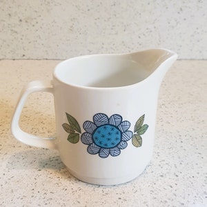 May include: White ceramic creamer with a blue and green floral design. The flower has a blue center with white stars and a white outline.