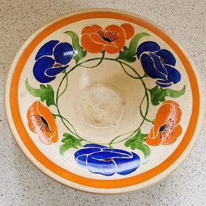 Può includere: Una ciotola in ceramica vintage con un motivo floreale. La ciotola presenta fiori blu e arancioni con foglie verdi su uno sfondo crema. La ciotola ha un ampio bordo arancione.