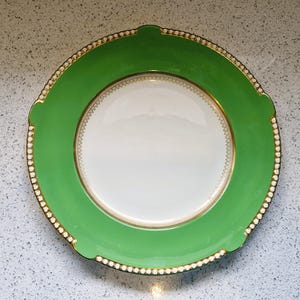 May include: A green and white ceramic dinner plate with a gold rim and a white centre. The plate has a decorative pattern around the edge.