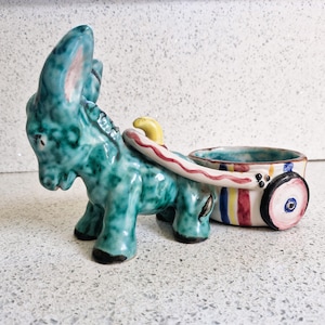 May include: A ceramic donkey figurine pulling a small, colorful cart. The donkey is teal blue with a pink nose and yellow hooves. The cart is white with red, yellow, and blue stripes and a pink and blue wheel.