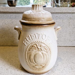Pode incluir: Um pote de cerâmica creme com tampa, com superfície texturizada e a palavra "RUMTOPF" em relevo. O pote tem um emblema decorativo de uma fruta com folhas. A tampa e a borda são marrons escuros. Duas alças estão nas laterais.