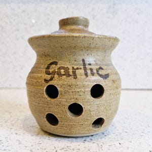 May include: A tan ceramic garlic keeper with a lid. The word "Garlic" is written on the front. The keeper has several round holes for ventilation. The keeper is a speckled tan color.