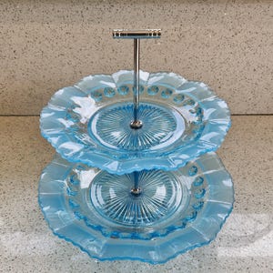May include: A two-tiered, light blue glass serving tray with a silver metal handle. The plates have a ruffled edge and a radial pattern in the center. The top plate is smaller than the bottom plate.