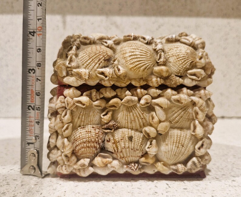 May include: A rectangular jewellery box, approximately 10 cm long, covered in various seashells. The box is beige and tan, with a red interior. The lid and sides are adorned with different sizes and shapes of shells.
