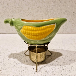 May include: A ceramic corn-shaped fondue pot with a green husk and yellow corn kernels. It sits on a small, gold-coloured metal stand with a fuel holder. The pot has a curved design and is likely used for serving melted cheese or chocolate.