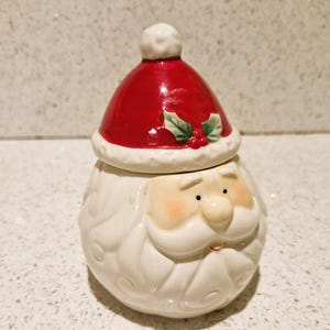 May include: White ceramic Santa Claus sugar bowl with a red hat and green holly. The bowl has a white glaze and a detailed face with a white beard.