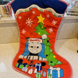 May include: Red Christmas stocking featuring Thomas the Tank Engine with a Santa hat, a green Christmas tree, snowflakes, and wrapped gifts. The stocking has a blue top and a red and white striped border.