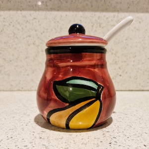 May include: A red ceramic jar with a white lid and a spoon. The jar has a painted design of a green leaf and a yellow pear.