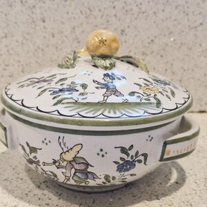 May include: A white ceramic serving dish with a lid. The dish is decorated with green and yellow floral designs and two figures in blue and yellow clothing. The lid has a yellow knob in the shape of a flower.