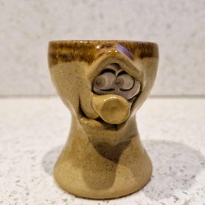 May include: A whimsical, handmade ceramic egg cup. The beige-colored cup features a face design with a surprised expression, complete with eyes and a protruding nose. The rim is a darker brown, adding contrast to the speckled surface.