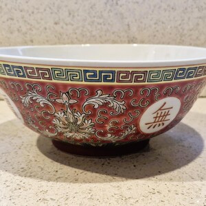 Peut inclure: Un bol en céramique décoratif avec un fond rouge et des motifs floraux blancs. Le bord présente un motif géométrique bleu, vert et jaune. Un ovale blanc avec un symbole marron est sur le côté. L'intérieur du bol est blanc.