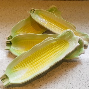 May include: Four ceramic corn on the cob serving dishes. The dishes are yellow and green and shaped like corn on the cob.
