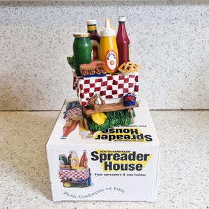 May include: A red and white checkered picnic table with a yellow mustard bottle, a red ketchup bottle, a green relish bottle, and a hot dog on a bun. The table is on a green base with a yellow banana and a red and white striped umbrella. The text on the box reads "Boston Warehouse Spreader House Four spreaders & one holder Picnic Condiments on Table".