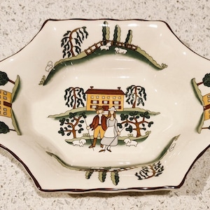 May include: A white ceramic serving dish with a brown rim and a green, yellow, and brown design. The design features a house, trees, and people.