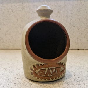 May include: A ceramic salt pig in a beige colour, featuring a brown rim and the word "SALT" embossed on the front. It has a large opening and a small knob on top. This kitchenware item is designed to hold salt for convenient use during cooking.