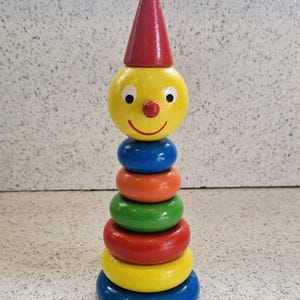 Könnte beinhalten: Ein Holz-Steckspielzeug in Form eines Clowns. Der Clown hat einen gelben Kopf mit roter Nase und roter Kegelhut. Der Körper besteht aus sechs farbigen Ringen: blau, orange, grün, rot, gelb und blau. Ein Spielzeug für Kinder.