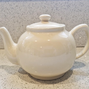 Op de afbeelding: Een witte theepot van keramiek met een rond lichaam en een tuit aan de linkerkant. De theepot heeft een deksel met een knop aan de bovenkant.