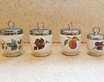 May include: Four white ceramic egg coddlers with silver lids. Each coddler features a different fruit or berry design: a single yellow rose, a cluster of red berries, a single peach, and a cluster of blackberries.