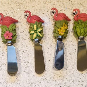 Peut inclure: Quatre couteaux à fromage décoratifs avec des figurines de flamants roses. Chaque couteau a une lame en acier inoxydable et un manche orné d'un flamant rose, de feuilles vertes et d'une fleur. Les couteaux sont disposés sur une surface mouchetée.