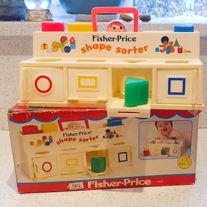 May include: A white plastic Fisher-Price shape sorter toy with a red handle. The toy has six different shaped holes and comes with a variety of colorful blocks. The box features a picture of a baby playing with the toy and the text "Fisher-Price shape sorter".