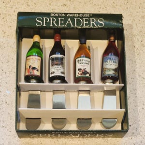 Puede incluir: Un juego de cuatro untadores en forma de botella de vino en miniatura en una caja verde y blanca. Los untadores están etiquetados como "Binding River", "Oakwood Cabernet", "Hill Flower Vineyard" y "Villa Pietra Valpolicella". La caja está etiquetada como "Boston Warehouse Spreaders".