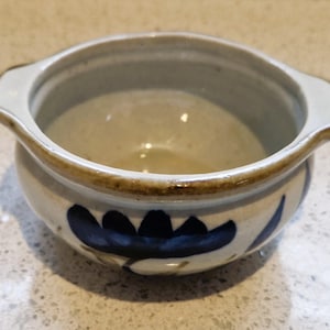 May include: A white ceramic bowl with blue and brown floral designs. The bowl has two handles on the sides and a slightly rounded shape.