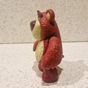 Peut inclure: Figurine d'ours rouge et beige avec une texture. L'ours a un nez brun fonc&eacute;, un ventre beige clair et une petite corne brun fonc&eacute;. La figurine est pos&eacute;e sur une surface blanche.