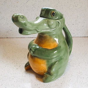 May include: A green and yellow ceramic teapot shaped like an alligator with a wide open mouth. The teapot has a lid and a handle.