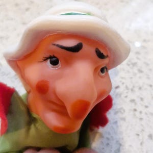 Puede incluir: Primer plano de una figura de gnomo vintage. La figura tiene una nariz grande, mejillas rosadas y un sombrero blanco con una banda verde. Viste una camisa verde con detalles rojos. El gnomo tiene cejas y pestañas negras.