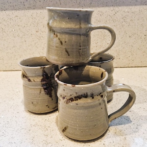 Puede incluir: Cuatro tazas de cerámica hechas a mano en tonos beige y marrón. Cada taza tiene un asa y un patrón moteado único. Las tazas están apiladas, mostrando su diseño rústico y artesanal.