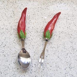Può includere: Un set di cucchiaio e forchetta con manici a forma di peperoncino rosso. Il cucchiaio e la forchetta sono in acciaio inossidabile e presentano un motivo decorativo a peperoncino con foglie verdi alla base. I peperoncini sono di colore rosso lucido.