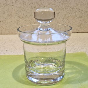 Puede incluir: Un tarro de vidrio transparente con una tapa redonda. El tarro está vacío y tiene una superficie lisa y pulida.