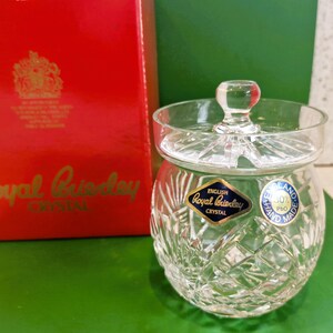 May include: A clear, cut-crystal jar with a lid, featuring a round knob. The jar has a diamond pattern and a blue and gold label that reads "Royal Brierley Crystal." A red box with gold lettering and a crest is in the background.