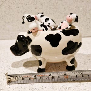 May include: A ceramic cow-shaped planter with a white base and black spots. The planter has four legs and contains several small cow figurines. The planter measures approximately 5 inches in length.
