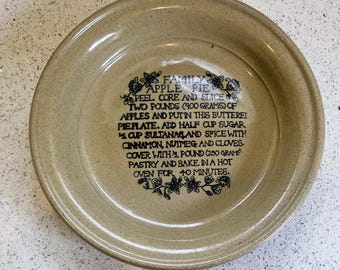 May include: A beige ceramic pie dish featuring a black printed apple pie recipe. The recipe specifies 900 grams of apples and other ingredients. The dish has a speckled finish and a slightly raised edge.