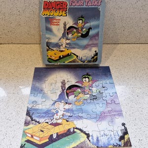 May include: A 50-piece Danger Mouse jigsaw puzzle featuring cartoon characters in a snowy landscape. The puzzle box is visible, displaying the title "Danger Mouse Four Tasks". The completed puzzle shows Danger Mouse and Penfold in a yellow car.
