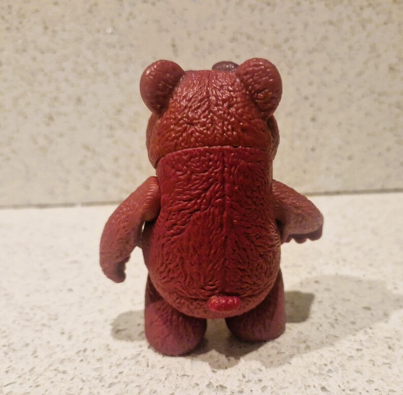 Peut inclure: Figurine d'ours en peluche de couleur marron, vue de dos. L'ours a une surface textur&eacute;e, de petites oreilles et des bras et des jambes trapus. La figurine est pos&eacute;e sur une surface de couleur claire.