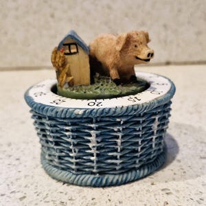 May include: A decorative kitchen timer featuring a light brown pig and a small building with a chicken figurine. The timer has a blue woven basket design and a white dial with black numbers.