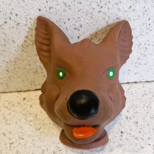 May include: A brown ceramic wolf head with green eyes and a black nose. The wolf's mouth is open, revealing an orange tongue. The sculpture has a detailed, textured surface and is likely a decorative item.