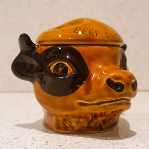 以下が含まれることがあります： 目の周りに黒い斑点があり、「ビーフドリップ」と書かれた蓋が付いた、茶色の陶器製の牛の頭の形をした容器です。