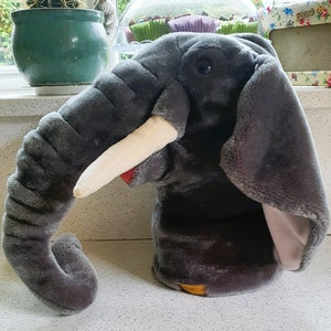 May include: Gray plush elephant hand puppet with a long trunk and large ears. The puppet has a white tusk and a red mouth. The puppet is sitting on a white surface.