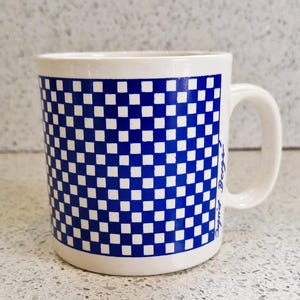 Może przedstawiać: Biały ceramiczny kubek z niebiesko-białym wzorem w szachownicę. Kubek ma zakrzywiony uchwyt, a na boku widoczny jest napis "Boyz". Kubek stoi na nakrapianej powierzchni.